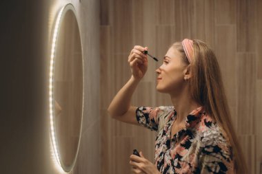 Sarışın kız aynanın önüne rimel sürüyor.