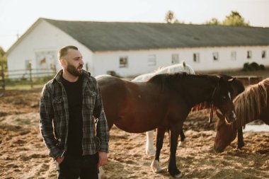 Genç bir çiftlik işçisinin portresi, arka planda atlarla birlikte. Genç bir çiftçi ahırda atlara bakar..