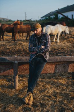 Genç bir çiftlik işçisinin portresi, arka planda atlarla birlikte. Genç bir çiftçi ahırda atlara bakar..