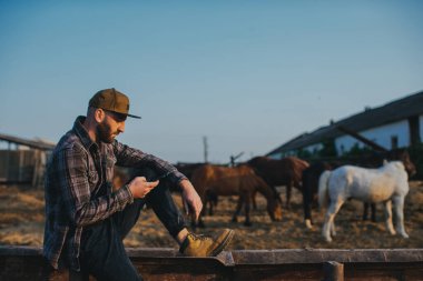 Elinde telefon olan genç bir adam atlarla bir otlağın arka planında. Bir adam çiftlikte bir günlük çalışmadan sonra dinlenir, gün batımında..