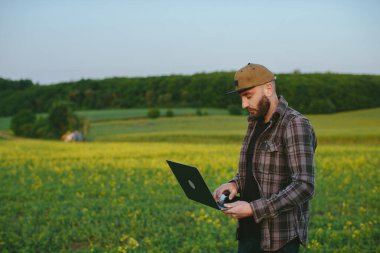 Bir çiftçi tarla işlerini denetler. Dizüstü bilgisayarı olan bir adam tarladaki bir traktörün çalışmasını izliyor..