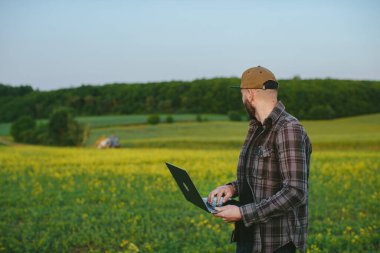 Bir çiftçi tarla işlerini denetler. Dizüstü bilgisayarı olan bir adam tarladaki bir traktörün çalışmasını izliyor..