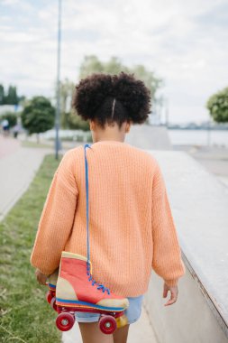 Genç bir kız kaykay yapmak için parka geldi. Afrikalı Amerikalı kız ayakkabı bağcıklarıyla bağlanmış eski püskü lastikler tutuyor..