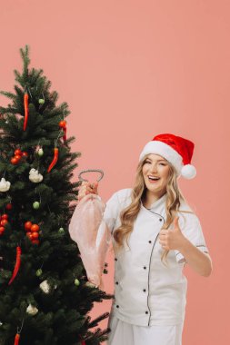 Yemek pişirme ve Noel tatili kavramı. Noel Baba şapkalı güzel sarışın bir aşçı Noel ağacının arka planında poz veriyor..