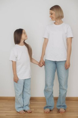 Anne ve küçük kız kot pantolon ve beyaz tişörtler giyip beyaz bir duvara poz veriyorlar..