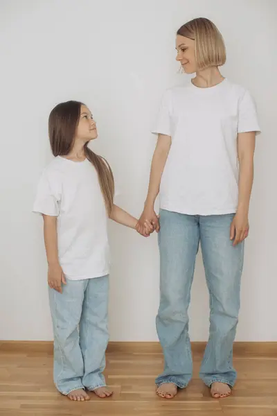 Mother and little daughter dressed in jeans and white t-shirts pose against a white wall.