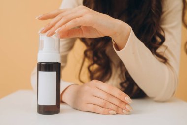 Fotoğraf maketi. Güzel, esmer bir kadının elinde bej bir arka planda izole edilmiş bir şişe kozmetik ürünle yakın çekimi..