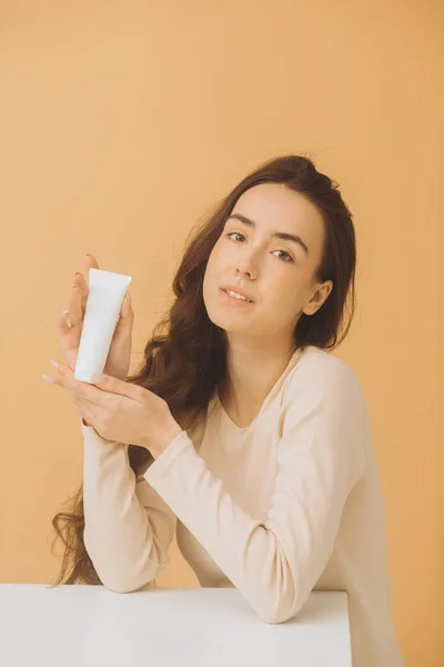A woman is holding a tube with cosmetic cream. Studio portrait of a pretty woman on a beige background.