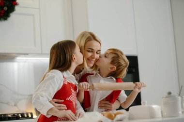 İki mutlu çocuk kış tatilinde evde Noel kurabiyesi yaparken annelerine sarılıyor. Mutlu anne, çocuklarıyla evde eğleniyor..