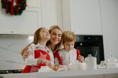 İki mutlu çocuk kış tatilinde evde Noel kurabiyesi yaparken annelerine sarılıyor. Mutlu anne, çocuklarıyla evde eğleniyor..