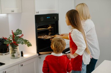 Anne ve çocuklar modern bir mutfakta Noel kurabiyesi pişiriyorlar. Annem tepsiyi kurabiyelerle birlikte fırına koyar. Aile tatili hazırlığı