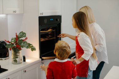 Anne ve çocuklar modern bir mutfakta Noel kurabiyesi pişiriyorlar. Annem tepsiyi kurabiyelerle birlikte fırına koyar. Aile tatili hazırlığı