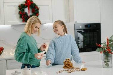 Mutlu anne ve tatlı kız Noel kurabiyelerini süslerken evde Noel süslemeleriyle sıcak mutfakta dikiliyorlar..