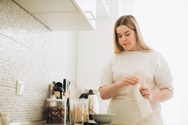 Mutfakta gülümseyen genç bir kadın, mısır gevreği ve sütten kahvaltı hazırlıyor..