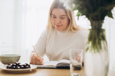 Güzel genç bir kadın mutfakta kahvaltı ederken günlüğüne not alıyor..