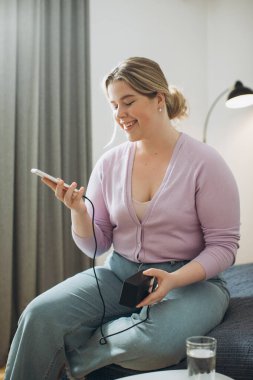 Genç bir kadın evdeki yatakta otururken telefonunu elektrik bankasıyla şarj ediyor..