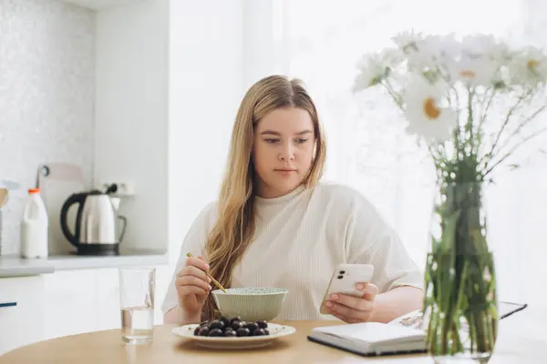 Den Blonda Flickan Äter Frukost Medan Hon Håller Telefonen Sina — Stockfoto