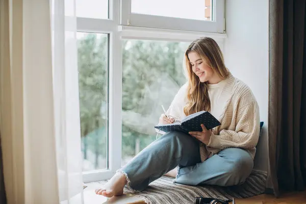 Creative young woman writes memory plan and idea in notebook while sitting near window at home. Creative journal or diary concept.