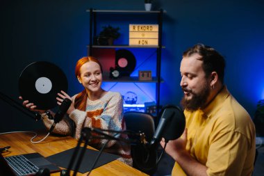 Müzik blogcuları podcast 'lerinin yeni bir bölümünü kaydediyor. Bir adam ve bir kadın stüdyoda müzik hakkında konuşuyorlar, kadın elinde plakla geziyor..
