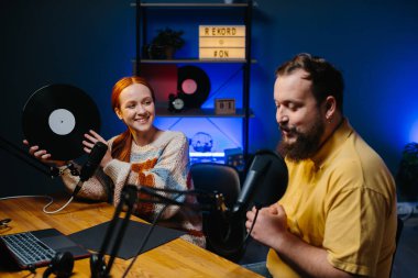 Müzik blogcuları podcast 'lerinin yeni bir bölümünü kaydediyor. Bir adam ve bir kadın stüdyoda müzik hakkında konuşuyorlar, kadın elinde plakla geziyor..