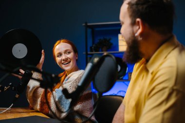 Müzik blogcuları podcast 'lerinin yeni bir bölümünü kaydediyor. Bir adam ve bir kadın stüdyoda müzik hakkında konuşuyorlar, kadın elinde plakla geziyor..