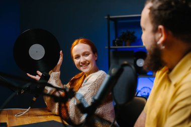 Müzik blogcuları podcast 'lerinin yeni bir bölümünü kaydediyor. Bir adam ve bir kadın stüdyoda müzik hakkında konuşuyorlar, kadın elinde plakla geziyor..