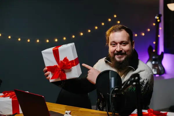ブロガーはチャンネルで視聴者にクリスマスプレゼントをオフにしています ホームスタジオでポーズしながら赤いリボンで縛られたギフトを持っている男 — ストック写真