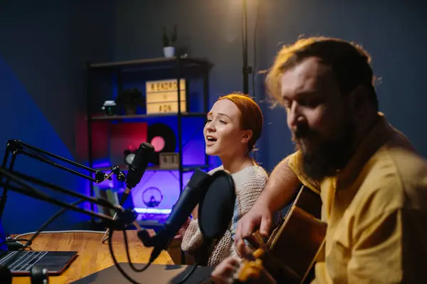 A cheerful couple of radio presenters perform a song on air. A man and a woman sing to the guitar in a home studio, creating content for their channel.