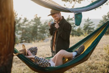 Çiftler dışarıda hamaklarda dinlenirken eğleniyorlar. Bir adam kamerada bir kızın fotoğrafını çekiyor..
