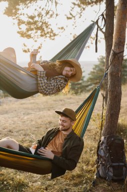 Bir erkek ve bir kız gün batımında bir çam ormanında hamaklarda dinleniyorlar.. 