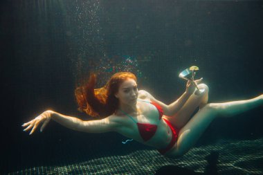 Güzel kızıl saçlı kadın yüzme havuzunda bir kokteyl ile poz veriyor. Yaz tatili kavramı.
