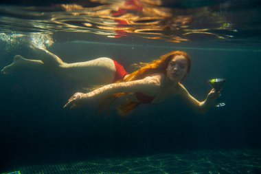 Güzel kızıl saçlı bir kadın suyun altında bir kokteyl ile poz veriyor..
