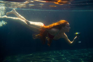 Güzel kızıl saçlı bir kadın suyun altında bir kokteyl ile poz veriyor..