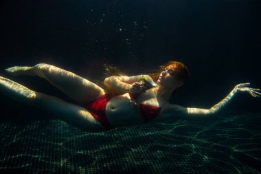 Güzel kızıl saçlı bir kadın suyun altında bir kokteyl ile poz veriyor..