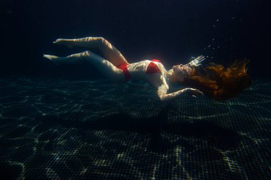 Güzel, sportif bir kız, yüzeyden gelen parlak güneş ışınlarına karşı gevşek saçlarıyla suyun altında poz verir..