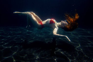 Güzel, sportif bir kız, yüzeyden gelen parlak güneş ışınlarına karşı gevşek saçlarıyla suyun altında poz verir..