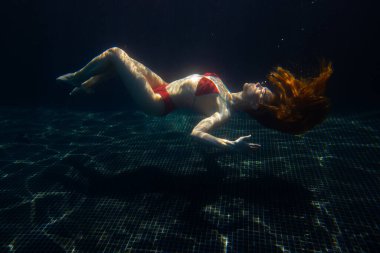 Güzel, sportif bir kız, yüzeyden gelen parlak güneş ışınlarına karşı gevşek saçlarıyla suyun altında poz verir..