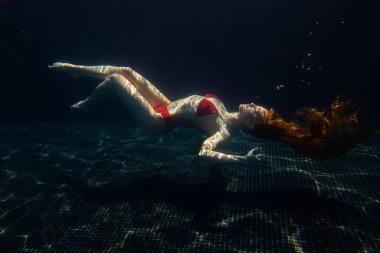 Güzel, sportif bir kız, yüzeyden gelen parlak güneş ışınlarına karşı gevşek saçlarıyla suyun altında poz verir..