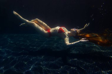 Güzel, sportif bir kız, yüzeyden gelen parlak güneş ışınlarına karşı gevşek saçlarıyla suyun altında poz verir..