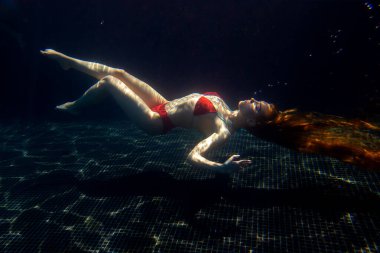 Güzel, sportif bir kız, yüzeyden gelen parlak güneş ışınlarına karşı gevşek saçlarıyla suyun altında poz verir..