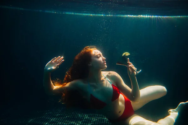 Beautiful red-haired woman posing with a cocktail underwater in a swimming pool. Summer vacation concept.