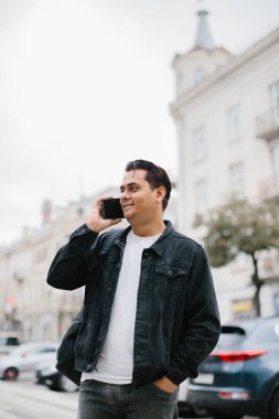 Genç Hintli bir adam sırt çantasıyla şehir caddesinde duruyor, cep telefonu kullanıyor, okuyor ve mesaj yazıyor, bir telefon görüşmesi yapıyor..