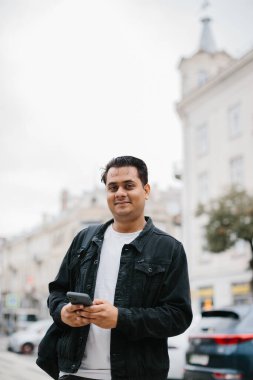 Genç Hintli bir adam sırt çantasıyla şehir caddesinde duruyor, cep telefonu kullanıyor, okuyor ve mesaj yazıyor, bir telefon görüşmesi yapıyor..