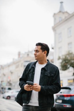 Genç Hintli bir adam sırt çantasıyla şehir caddesinde duruyor, cep telefonu kullanıyor, okuyor ve mesaj yazıyor, bir telefon görüşmesi yapıyor..
