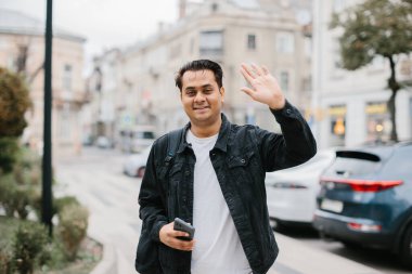Mutlu gülümseyen Hintli turist elinde telefonla merhaba diyor. Dost canlısı bir adam bir şehir caddesinde dururken elini sallıyor..