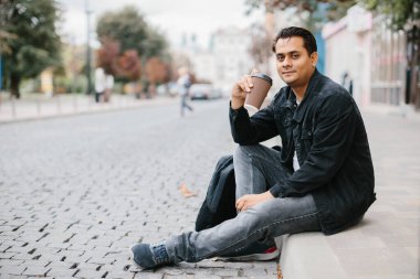 Gülümseyen bir Hindu adam bir şehir caddesinde yerde oturuyor ve sabah kahvesinin tadını çıkarıyor. Turizm ve şehir yaşam tarzı kavramı.