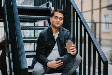 Genç bir Hindu adam şehir caddesinde metal bir merdivende oturuyor ve telefonunu kontrol ederken sabah kahvesinin tadını çıkarıyor. Turizm ve şehir yaşam tarzı kavramı.