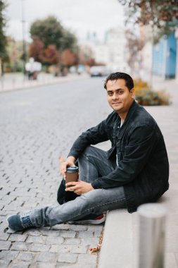 Gülümseyen bir Hindu adam bir şehir caddesinde yerde oturuyor ve sabah kahvesinin tadını çıkarıyor. Turizm ve şehir yaşam tarzı kavramı.
