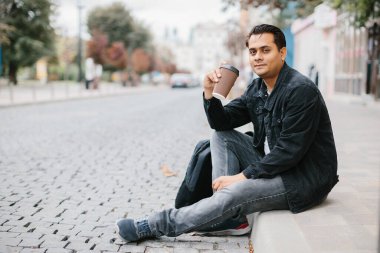 Gülümseyen bir Hindu adam bir şehir caddesinde yerde oturuyor ve sabah kahvesinin tadını çıkarıyor. Turizm ve şehir yaşam tarzı kavramı.