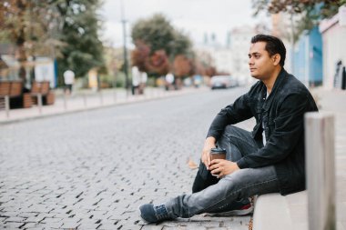 Gülümseyen bir Hindu adam bir şehir caddesinde yerde oturuyor ve sabah kahvesinin tadını çıkarıyor. Turizm ve şehir yaşam tarzı kavramı.